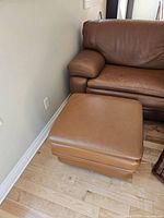 Tan Italian leather ottoman placed beside a matching brown leather sofa on a light wooden floor, showing clean condition and rectangular shape.