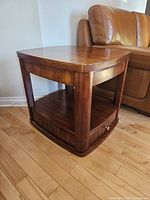 Side angle view of the Mazin wooden side table showing the drawer pulled out, open lower shelf, legs, and tabletop surface next to a leather couch on hardwood flooring.