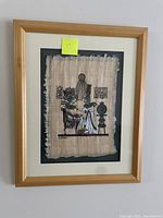 Framed Egyptian papyrus artwork hanging on a white wall, showing two seated figures and Egyptian hieroglyphs on the papyrus inside a wood-colored frame with cream matte.