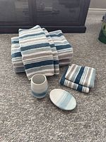 Photo shows matching set of six towels (bath, hand, face cloth) in stripes of beige, blue, white alongside ceramic soap dish and ceramic cup with gradient blue and white.