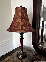 Full view of the lamp showing red gold brocade fabric shade and the lamp base on a dark surface next to a mirror.