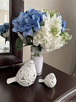 Blue and white artificial flower arrangement in a clear glass vase with a rhinestone band near the top, placed on a dark wood surface with two white ceramic decorative birds in front.