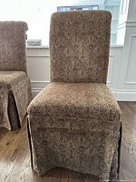 Two Bombay Co. parson chairs upholstered in patterned fabric with skirts covering the legs and tassels on the back, positioned on a wooden floor near white cabinetry.