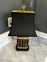 Front view of lamp showing black base with gold Chinese calligraphy on each side and black pleated lampshade with gold interior trim and decorative finial on top.