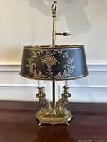 Front view of the lamp showing black painted tole shade with gold floral decoration, weighted brass base with two brass dolphin candle holders.