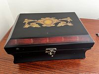 Top angled view of the black wooden jewelry chest showing the inlaid gold and brown decorative pattern on the lid and curved base.