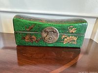 Front view of a green wooden box featuring gold floral and bird artwork and a round metal lock plate