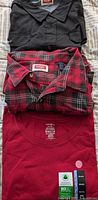 Three folded shirts showing black Wrangler shirt on top, red and black Wrangler plaid shirt in middle, and red sleeveless T-shirt on bottom with tags visible