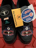 Top view of pair of black men's safety shoes with CSA Grade 1 steel toe and steel plate certification tags, size 8 labels on shoes, and slip resistant sole tag.