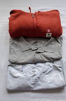 Three men's tops folded and stacked: rust zipper hoodie, gray polo shirt, white t-shirt