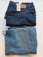 Two folded pairs of brand new women's denim jeans, one dark blue with Signature for Wrangler label and one medium blue, displayed on white surface.