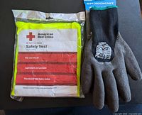 Photo showing American Red Cross safety vest in original sealed packaging next to one black and grey thermal lined work glove with grip