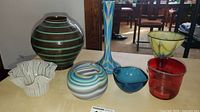 Photo showing all seven glass pieces on a table with brown vase in back and other colorful bowls and vases in front