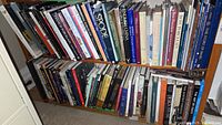 Wide view of two shelves filled with numerous porcelain, pottery, and glass art books in dark and light covers, stacked upright and side by side.
