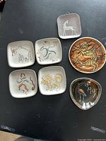 Photo showing all 7 ceramic items: 4 square plates with hand-painted designs and 3 other decorative dishes of varying shapes and colors placed on black surface.