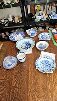 Overall set of Delft style blue and white ceramic cups, plates, bowls, and utensils shown on a wooden table with shelving in the background.