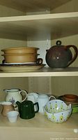 Top and middle shelves showing tan shallow bowls, brown/green glazed pitcher with lid, assorted small creamers and cups, stacked colorful dipping bowls.