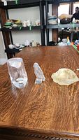 Three glass art objects displayed on a wooden table including a teardrop bowl with face design, a bird figurine, and a shell-shaped dish.
