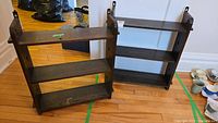 Pair of two vintage wood display shelves standing on a wood floor. Both have three shelves, dark wood look, with mounting brackets on sides.