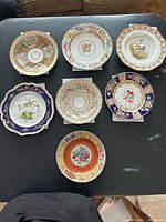 Top-down view of the seven assorted porcelain plates arranged on a black surface showing varied floral and decorative designs in multiple colors with some plates having gold accents and scalloped edges.