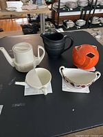 Side view of all five pottery and porcelain items on table showing black pitcher, orange vase, white teapot, small white pot, porcelain dish.