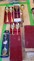 Lot of souvenir spoons and a pewter plate shown with most items laid out on a surface with green tape