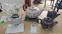 Group of six teapots and one green trivet on tabletop