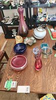 Group shot of all lot items on wooden table including glass vases, bowls, dishes, paperweights, and lidded ceramic jar