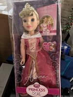 Photo of Aurora doll in original packaging showing detailed pink and gold ball gown, tiara, and accessories visible through box.