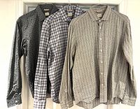 Three men's cotton button-down shirts on hangers: dark grey with deer print, light beige check, black and white grid check.