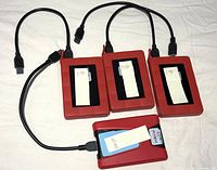 Four external hard drives arranged on a white surface, three labeled 1TB and one labeled 500GB. Each connected to a black USB 3.0 cable. All have red housings except one with identifiable blue label area.