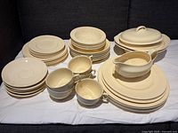 Full set displayed showing plates, cups, bowls, serving dishes and gravy pourer on white cloth and grey sofa.