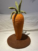 Photo of an unused carrot-shaped cat scratching post standing on a white cloth background. The post is 17 inches tall, orange rope wrapped body, green plush leaves on top, on a brown circular base.