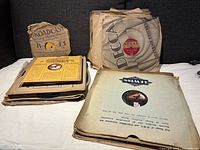 Stack of vintage 78 rpm records in original paper sleeves from labels including Decca, Regal, and Lewis, arranged on a couch showing paper sleeve wear and vintage label designs.