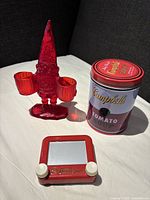 Full lot view with red acrylic garden gnome organizer holding two baskets next to Campbell's Tomato Soup puzzle tin and red Pocket Etch A Sketch on light surface.