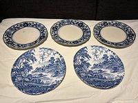 Five vintage plates laid out on a flat surface, showing two plates with detailed blue countryside scenes and three plates with blue floral decorated rims on cream background.
