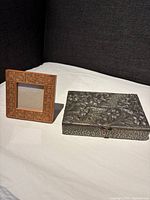 Photo showing the pewter flower-embossed box and the carved wooden picture frame side by side on a white surface with a dark background.