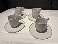 Four light grey porcelain coffee cups and saucers arranged on a cloth, showing front angled view of set with square cup handles and matching round saucers.