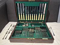 Full view of opened wooden cutlery box lined with green felt, containing knives, forks, spoons, ladles, and napkin rings, arranged neatly in compartments.