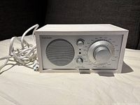 Front view of the white Tivoli Audio Henry Kloss Model One radio/speaker showing speaker grill, tuning knob, and volume knob with power cable.