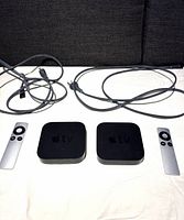 Front view of two Apple TV 3rd generation units with their corresponding aluminum remotes and power cables on a white surface