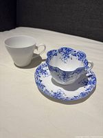 Photo showing two china teacups, a scalloped blue and white floral Shelley Dainty Blue teacup with matching saucer, in front of a plain white Rosenthal Studio-Line teacup without saucer.