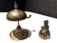 Front view of vintage brass reception bell, about 4 inches tall with a rounded top and lever, next to a smaller dinner/service bell shaped like a figurine of a matron.