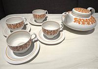 Teapot and four matching teacups on saucers arranged on white surface, showing orange decorative pattern.