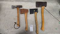Overview of three small axes and one wood splitter arranged on floor, showing wooden handles and metal heads. Two small axes have leather sheaths, one handle has leather strap loop.
