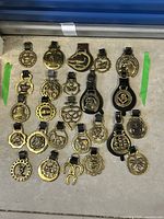 Overview photo showing the collection of 23 brass horse medallions laid out flat, most attached to leather straps.