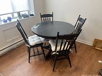 Full view of round black wood breakfast table with 4 matching spindle-back chairs with white cushions in a room with wood floor and window.
