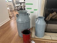 Front view of large Grahams Creamery Co. milk can, Modern Dairies milk can, and small red tin jug grouped together on hardwood floor near fireplace.