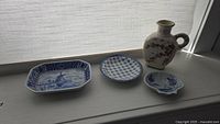 Four ceramic and stoneware items arranged on a window ledge: rectangular Delft plate, round blue and white plate, small shell shaped bowl, stoneware jug with floral design.