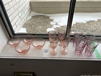 4 pink swirled glass bowls, 4 matching pink swirled stemmed glasses, and 4 rose-colored textured glass tumblers arranged on a windowsill.